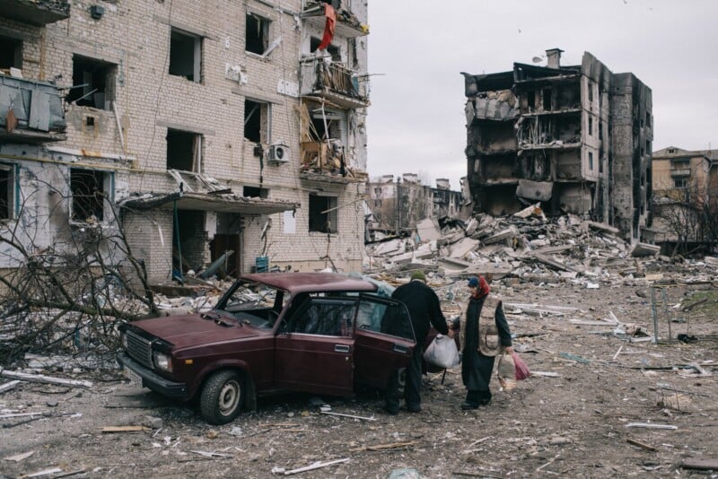 Two people with bags stand beside a maroon car in front of heavily damaged, partially collapsed apartment buildings in a debris-strewn area, suggesting recent destruction or conflict.