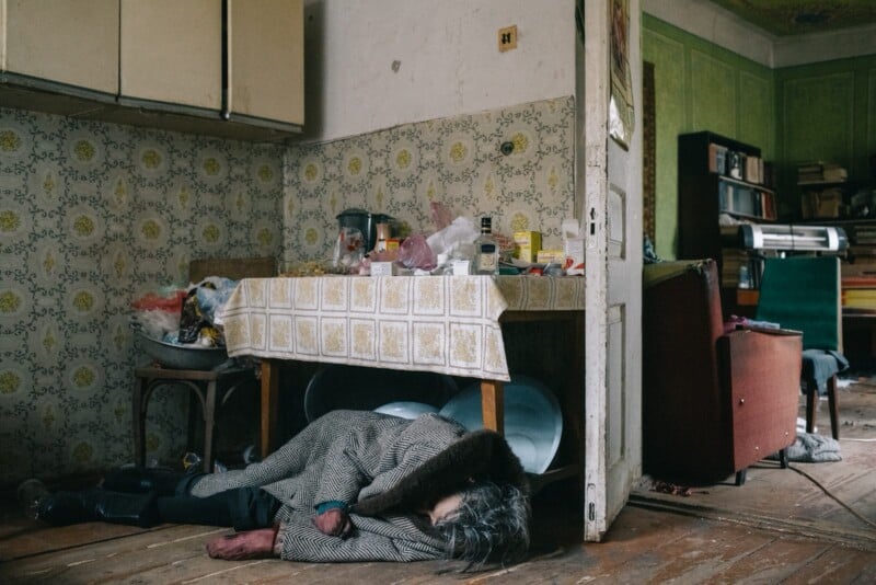 An elderly person lies on the floor under a table in a cluttered, dimly lit kitchen with peeling wallpaper and scattered household items, suggesting a state of distress or neglect.