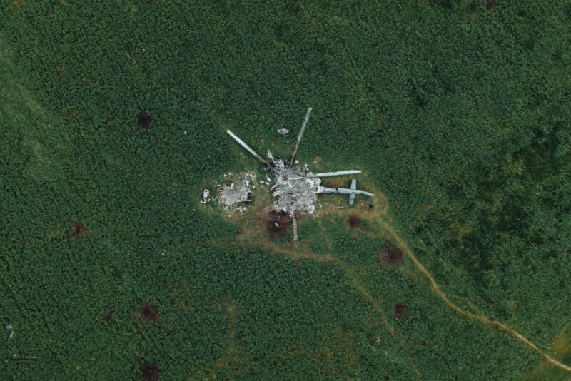 Aerial view of a helicopter crash site in a green field, with debris and helicopter parts scattered in a circular pattern around the impact area.