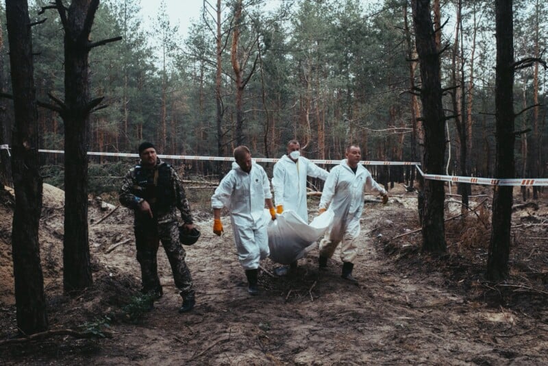 Four people in white hazmat suits carry a body bag through a forested area, while another person in camouflage gear walks beside them. Police tape is visible among the trees in the background.