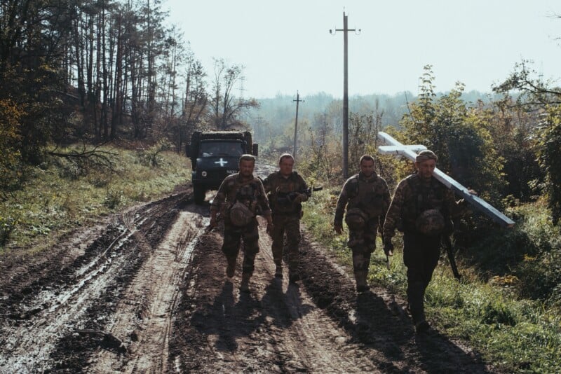 Four soldiers in camouflage walk along a muddy dirt road carrying a large white cross, with a military truck and trees in the background under a bright sky.