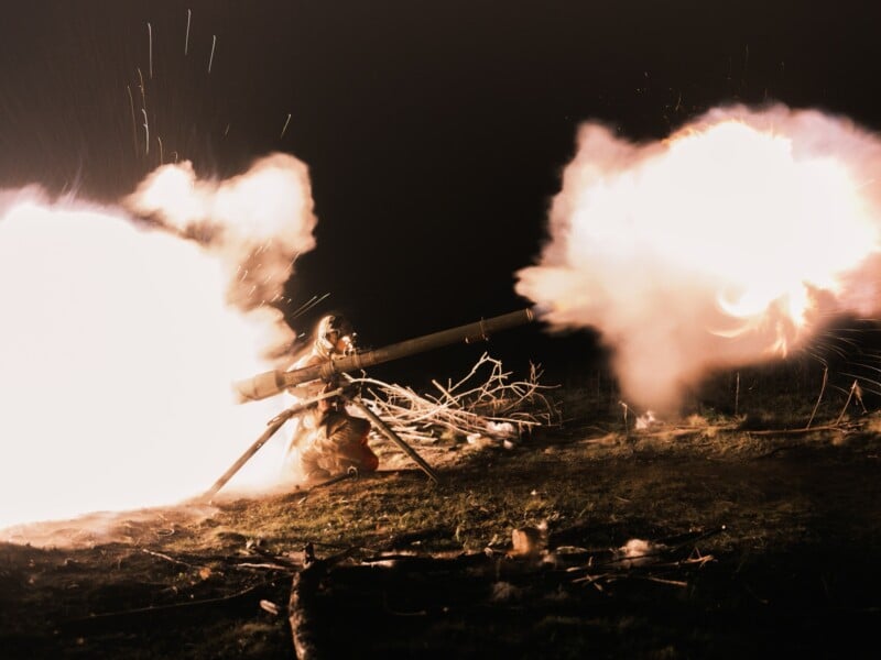 A person firing a large, tripod-mounted weapon at night, producing a bright flash and cloud of smoke from the barrel. The intense light illuminates the ground and nearby branches.