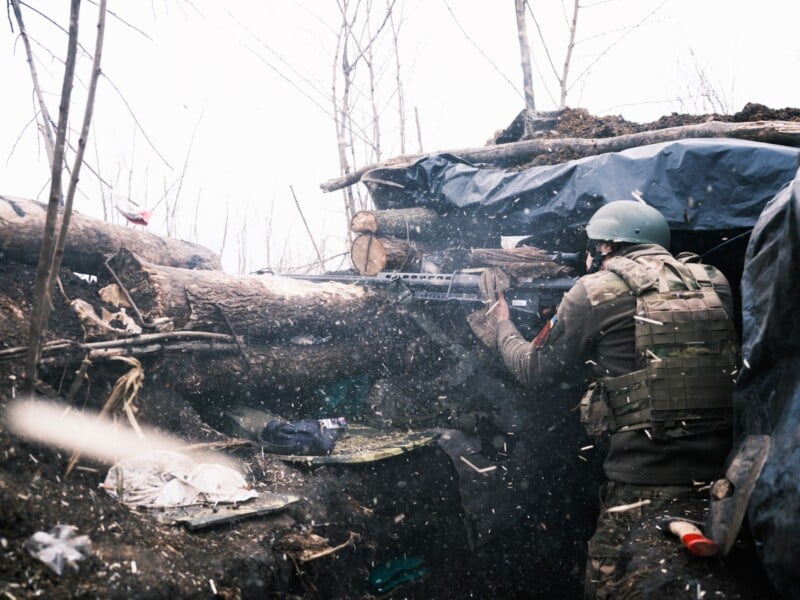 A soldier in camouflage gear and helmet fires a weapon from behind a barricade of logs and dirt in a trench, with debris and splinters flying in the air, surrounded by leafless trees.