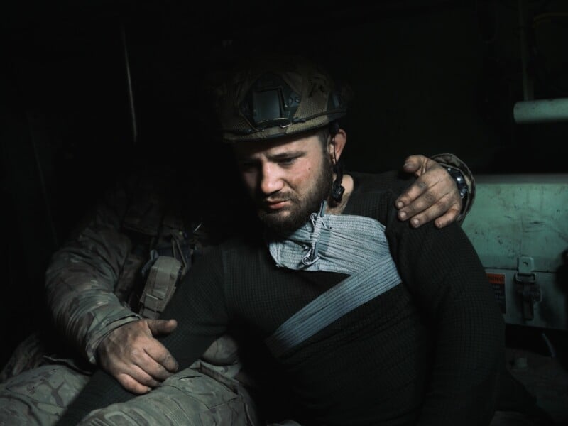 A soldier wearing a helmet and dark clothing sits with a bandaged shoulder, looking down somberly as another person, mostly in shadow, rests a comforting hand on his arm. The scene is dimly lit, suggesting a tense atmosphere.