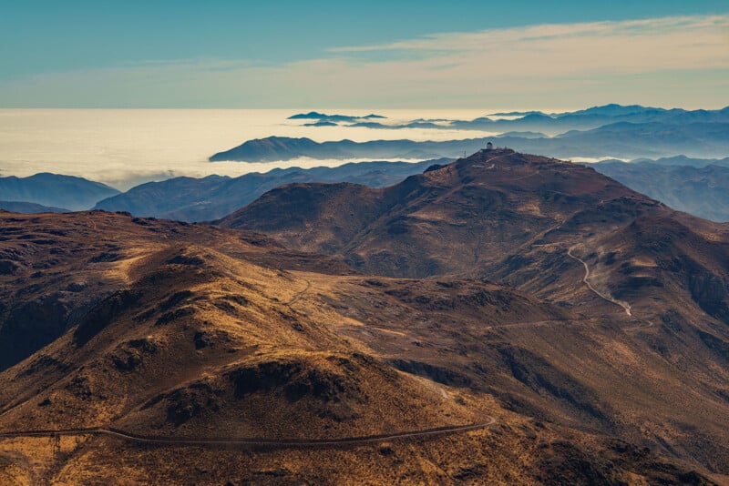 A panoramic view of rugged, brown mountains under a blue sky, with winding roads and a distant observatory on one peak. In the background, clouds hover low above distant mountains and the horizon.