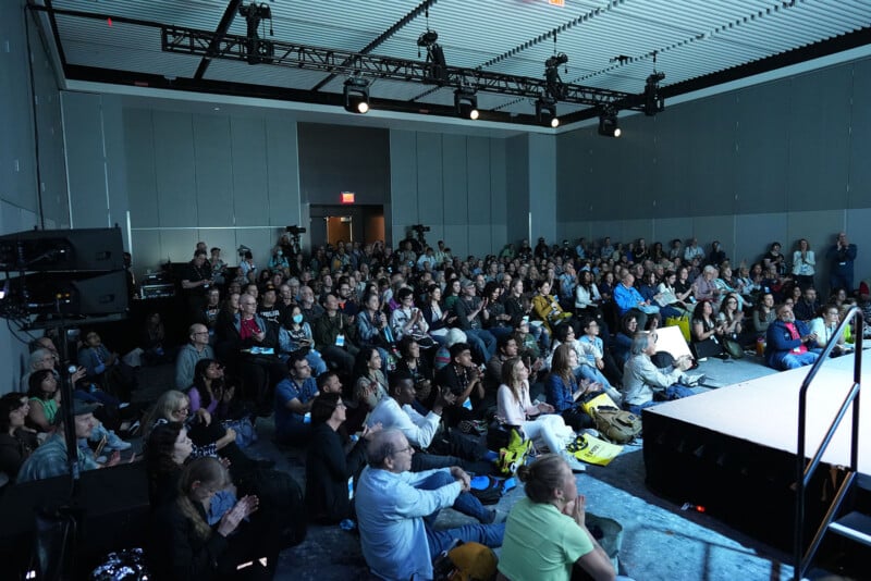 A large audience sits attentively in a conference room, facing a stage at the front. The room is well-lit, with rows of chairs filled with people of diverse backgrounds. Some are taking notes or holding event bags.