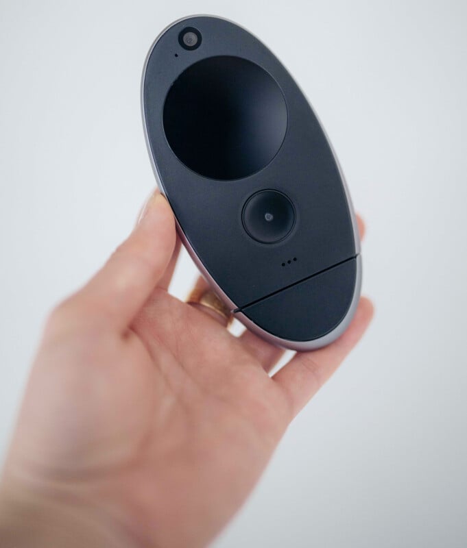 Kyu Camera Unveiled: Revolutionizing the Art of Memory Capture 13 A hand holding a small, oval-shaped black electronic device with a camera lens and a circular touchpad on its surface against a plain light background.