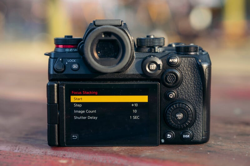 A digital camera on a wooden surface with its rear screen displaying focus stacking settings, including step, image count, and shutter delay options. The camera is outdoors with a blurred background.