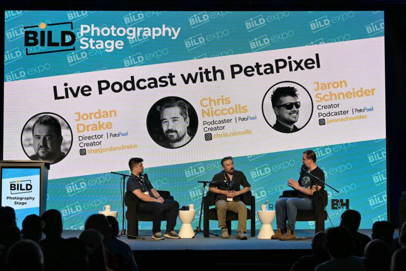 Three men sit on stage with microphones, engaged in a panel discussion. Behind them, a large screen reads, "Live Podcast with PetaPixel" and displays portraits and names: Jordan Drake, Chris Niccolls, and Jaron Schneider.