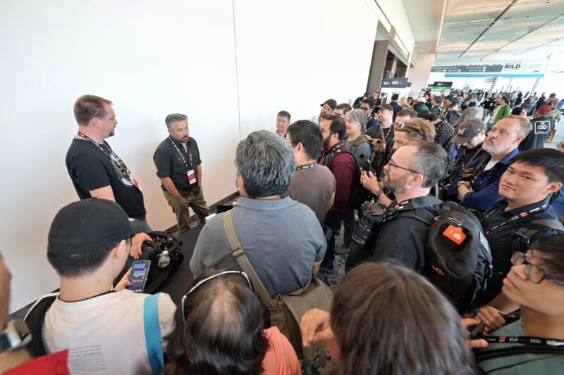 A large group of people gathers closely around two men speaking at a table in a convention hall. Several attendees hold cameras or wear camera gear, and event signage is visible in the background.