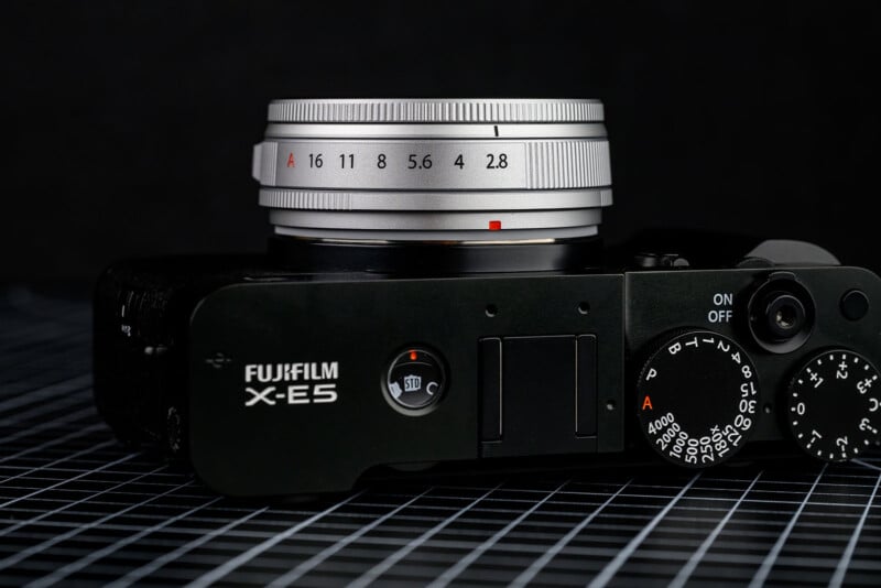 Close-up of the top view of a Fujifilm X-E5 camera with a silver lens, showing aperture settings and control dials, placed on a black grid-patterned surface.