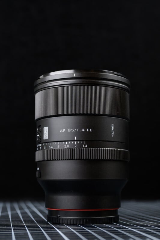 A black Viltrox AF 85mm f/1.4 FE camera lens stands upright on a grid-patterned surface, shown against a dark, textured background.