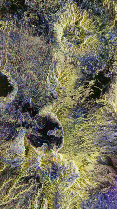 Satellite image showing a textured, colorful landscape with circular, crater-like formations, ridges, and valleys in shades of green, yellow, purple, and black. Patterns suggest volcanic or erosion features.