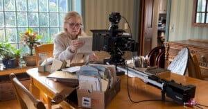 A woman sits at a wooden table examining documents, surrounded by boxes of papers. A camera on a slider is positioned in front of her, and sunlight streams in from a window with houseplants on the sill.