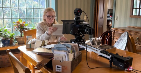 A woman sits at a wooden table examining documents, surrounded by boxes of papers. A camera on a slider is positioned in front of her, and sunlight streams in from a window with houseplants on the sill.