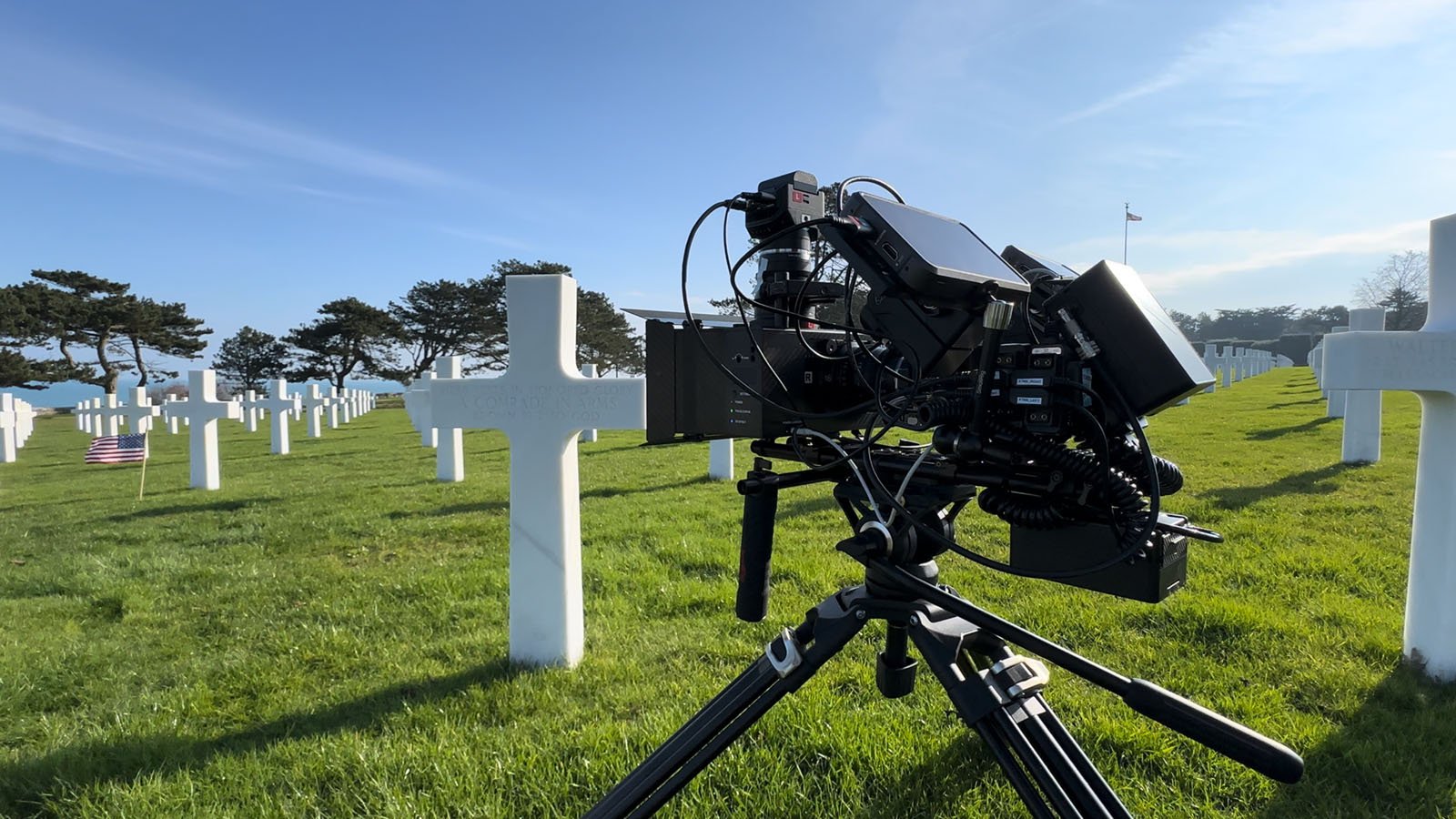 A professional video camera on a tripod is set up in a cemetery with white crosses on green grass under a clear blue sky. Some gravestones have small American flags placed in front of them.
