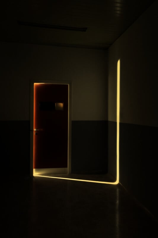 A dimly lit hallway with a closed door, illuminated by a bright, narrow beam of light streaming through the door's edge and casting a sharp L-shaped line on the dark wall and floor.