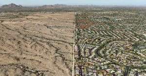 Aerial view showing a stark divide between a dry, barren desert landscape on the left and a densely populated suburban neighborhood with houses, greenery, and winding streets on the right.
