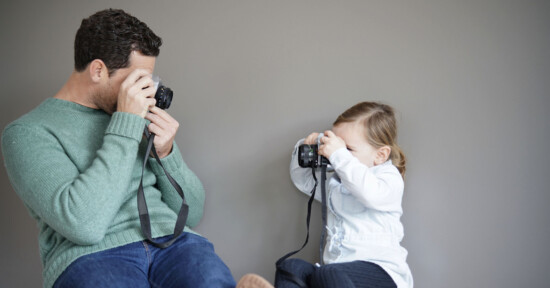 A man and a young girl sit next to each other, both playfully holding cameras up to their faces as if taking photos of each other against a plain gray background.