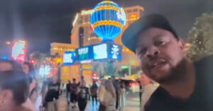 A man in a black cap poses in the foreground of a lively, neon-lit street at night, with the illuminated Paris Las Vegas balloon sign and crowds of people visible in the background.