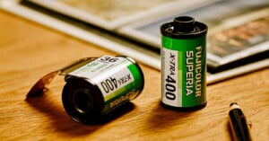 Two Fujicolor Superia X-TRA 400 film canisters, one standing upright and one lying on its side, are placed on a wooden surface near a pen and a photo album.
