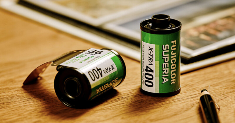 Two Fujicolor Superia X-TRA 400 film canisters, one standing upright and one lying on its side, are placed on a wooden surface near a pen and a photo album.