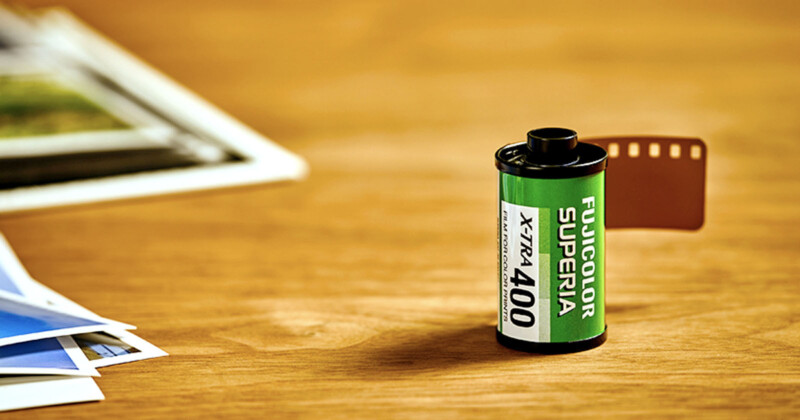 A Fujicolor Superia X-TRA 400 film canister sits on a wooden surface, with a few printed photos blurred in the background. A small piece of brown film is protruding from the canister.