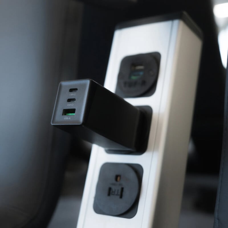 A close-up of a power strip with multiple outlets, including a black USB charger plugged into one socket and additional USB ports and power outlets visible. The background is slightly blurred.