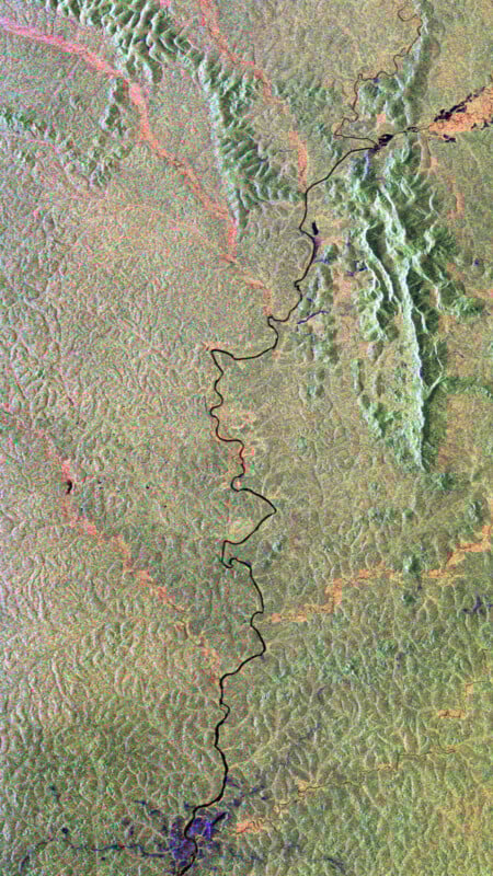 A satellite view of a winding river cutting through a mostly green and brown landscape, with textured ridges and patches of vegetation visible throughout the image.