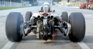 A rear view of a vintage Formula 1 race car on a track, showing wide tires, exposed suspension, and engine components, with some smoke rising from the left tire.
