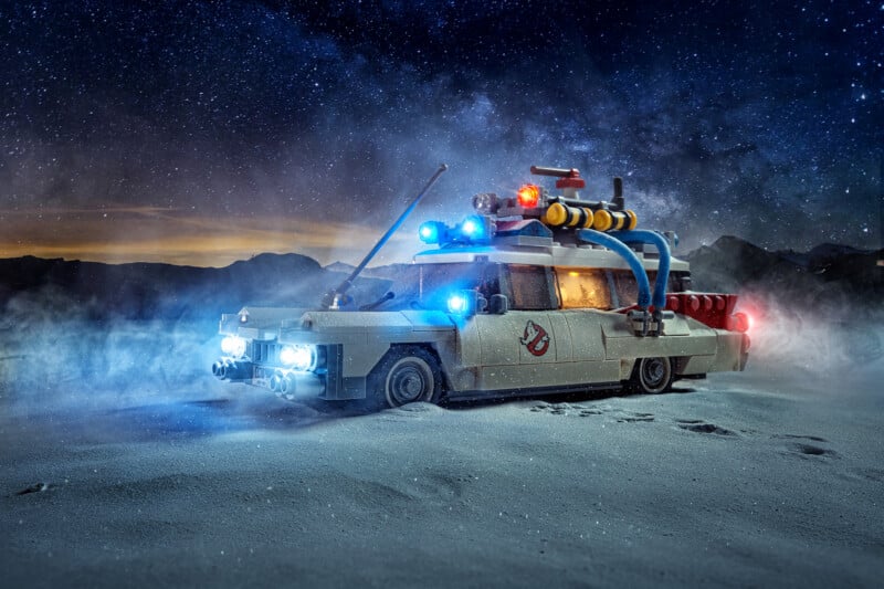 A LEGO model of the Ghostbusters Ecto-1 car with glowing headlights and rooftop lights sits on a snowy landscape under a starry night sky, surrounded by mist.