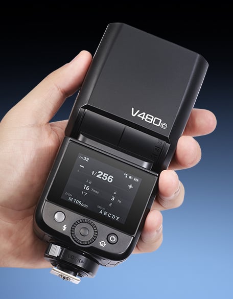 A hand holding a black V480C camera flash unit, displaying its digital control panel and settings on the screen against a gradient blue background.