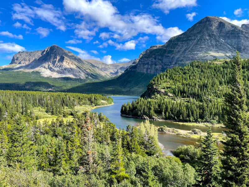 A scenic view of a river winding through a lush green forest, with tall mountains in the background under a bright blue sky with scattered clouds.