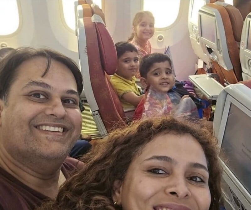 A family of five—two adults and three children—smile for a selfie while sitting in their seats on an airplane. The cabin and seatback screens are visible in the background.