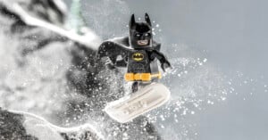A LEGO Batman minifigure snowboarding down a snowy slope, with snow flying around him and a determined expression on his face.