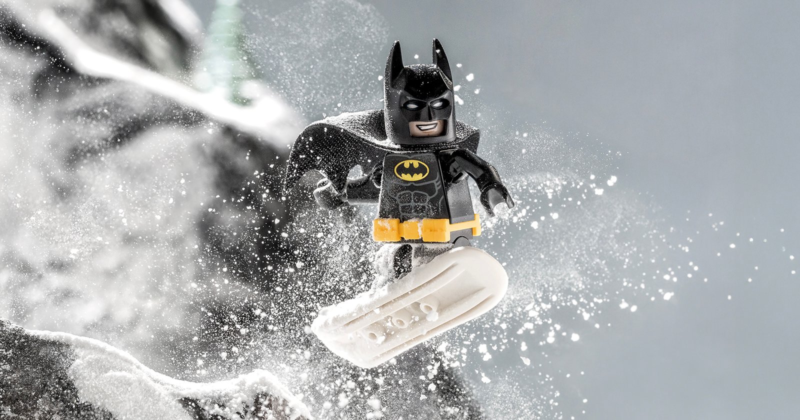 A LEGO Batman minifigure snowboarding down a snowy slope, with snow flying around him and a determined expression on his face.
