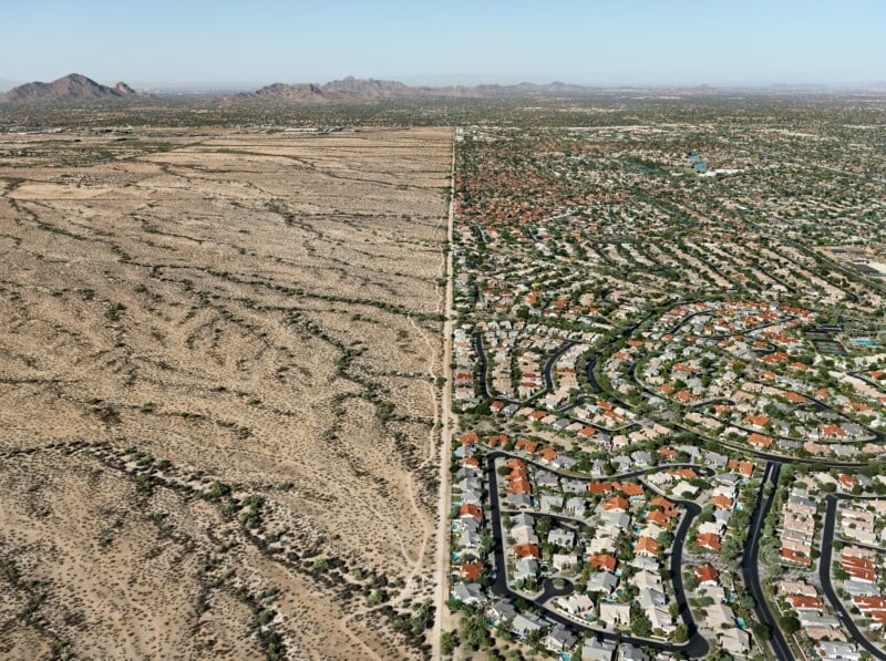 Aerial view showing a sharp boundary between desert landscape on the left and a large suburban neighborhood with houses, green lawns, and winding roads on the right; mountains visible in the distance.