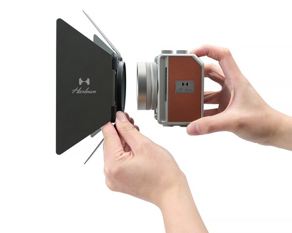 A person’s hands attach a rectangular lens hood to a vintage-style camera with a brown leather grip and a silver lens. The camera and hood both display the “Herklove” logo.