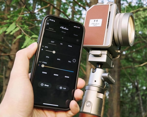 A hand holds a smartphone displaying lighting control settings next to a vintage-style camera or light mounted on a handheld stabilizer, with a forest background.