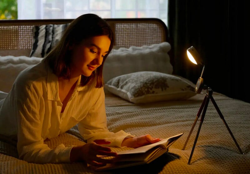 A woman in a white shirt lies on a bed, reading a book by the light of a small lamp on a tripod. The room is softly lit with a cozy atmosphere, and the bed has patterned pillows and a cushioned headboard.