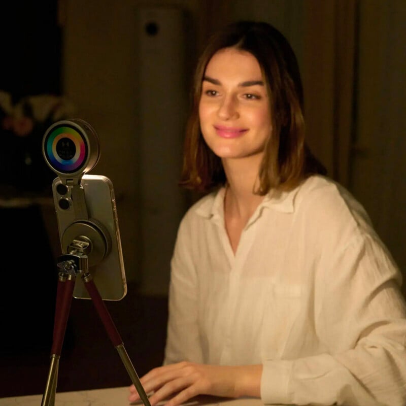 A woman in a white blouse sits at a desk, smiling at a smartphone mounted on a tripod with a colorful ring light in a dimly lit room.