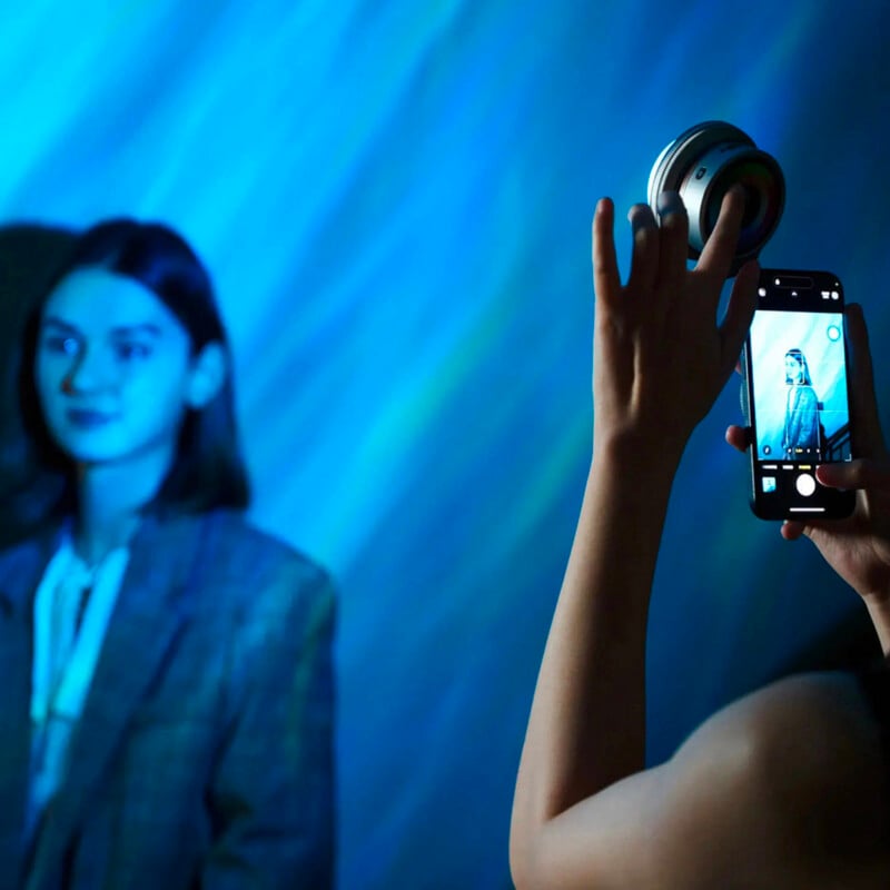 A person holding a smartphone and circular light takes a photo of a woman standing against a blue, softly lit background. The woman has shoulder-length hair and wears a checked blazer.