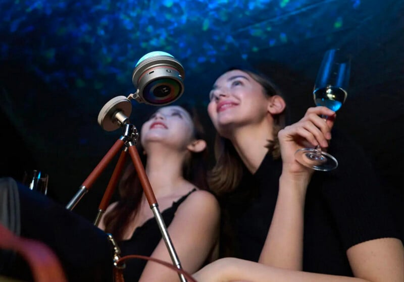 Two women sit close together, smiling and looking up at a starry projection. One holds a glass of wine, while a device on a tripod projects the star-like lights onto the dark ceiling.