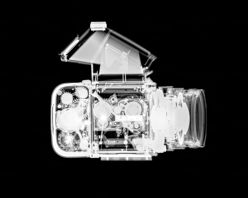 X-ray image of a camera, showing internal gears, mechanisms, and lens elements in white against a black background. The camera’s viewfinder is open, revealing intricate details inside.