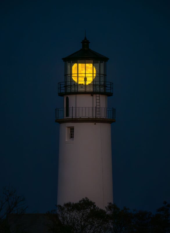 A full moon appears perfectly aligned inside the top of a tall white lighthouse against a dark evening sky, creating the illusion that the moon is the lighthouse’s glowing light.