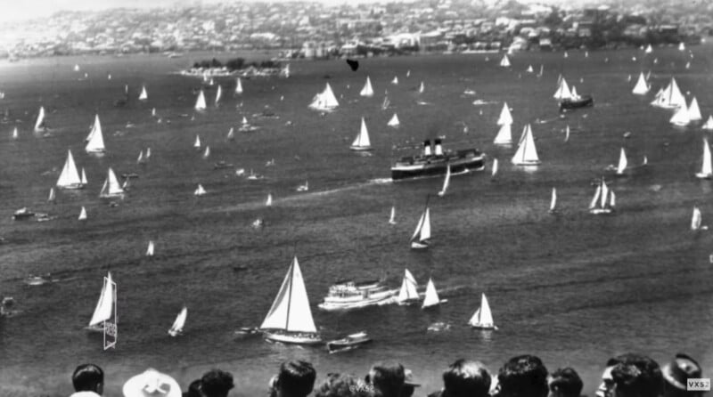 Black-and-white photo of a busy harbor filled with sailboats and a large ship. People watch the scene from a hillside in the foreground, with a cityscape visible across the water in the background.