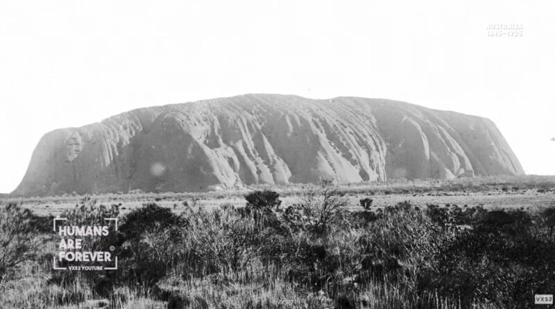 Black and white photo of Uluru, a large rock formation in Australia, seen from a distance with vegetation in the foreground. Text on the image reads “HUMANS ARE FOREVER” and “AUSTRALIA 146-17525.”.