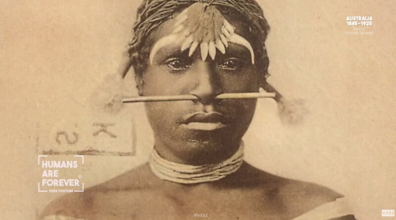 A sepia-toned portrait of an Indigenous Australian person with traditional facial adornments, including a horizontal nose piercing and feathered decorations. The person wears a woven headpiece and multiple necklaces.