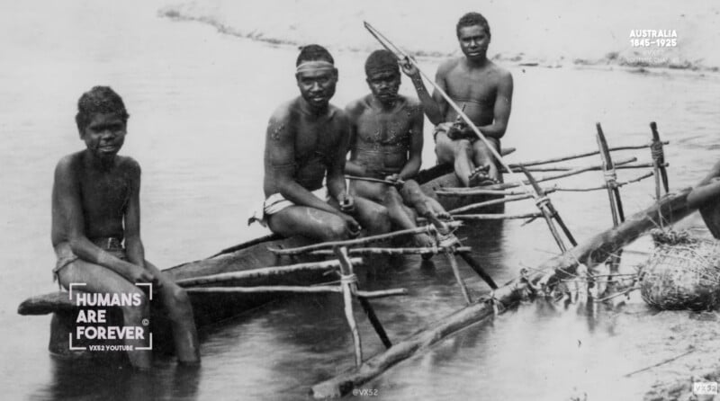 Four Indigenous Australian men sit on wooden canoes with outriggers in shallow water near a sandy bank. They are minimally clothed and looking toward the camera. The photo has a "Humans Are Forever" watermark.
