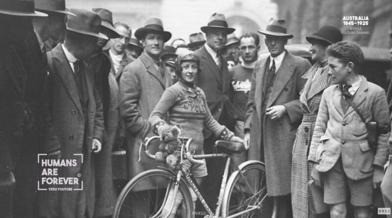 A woman in vintage cycling gear stands beside her bicycle, surrounded by a group of men in suits and hats. The photo appears to be from the early 20th century, and the crowd looks on with interest.